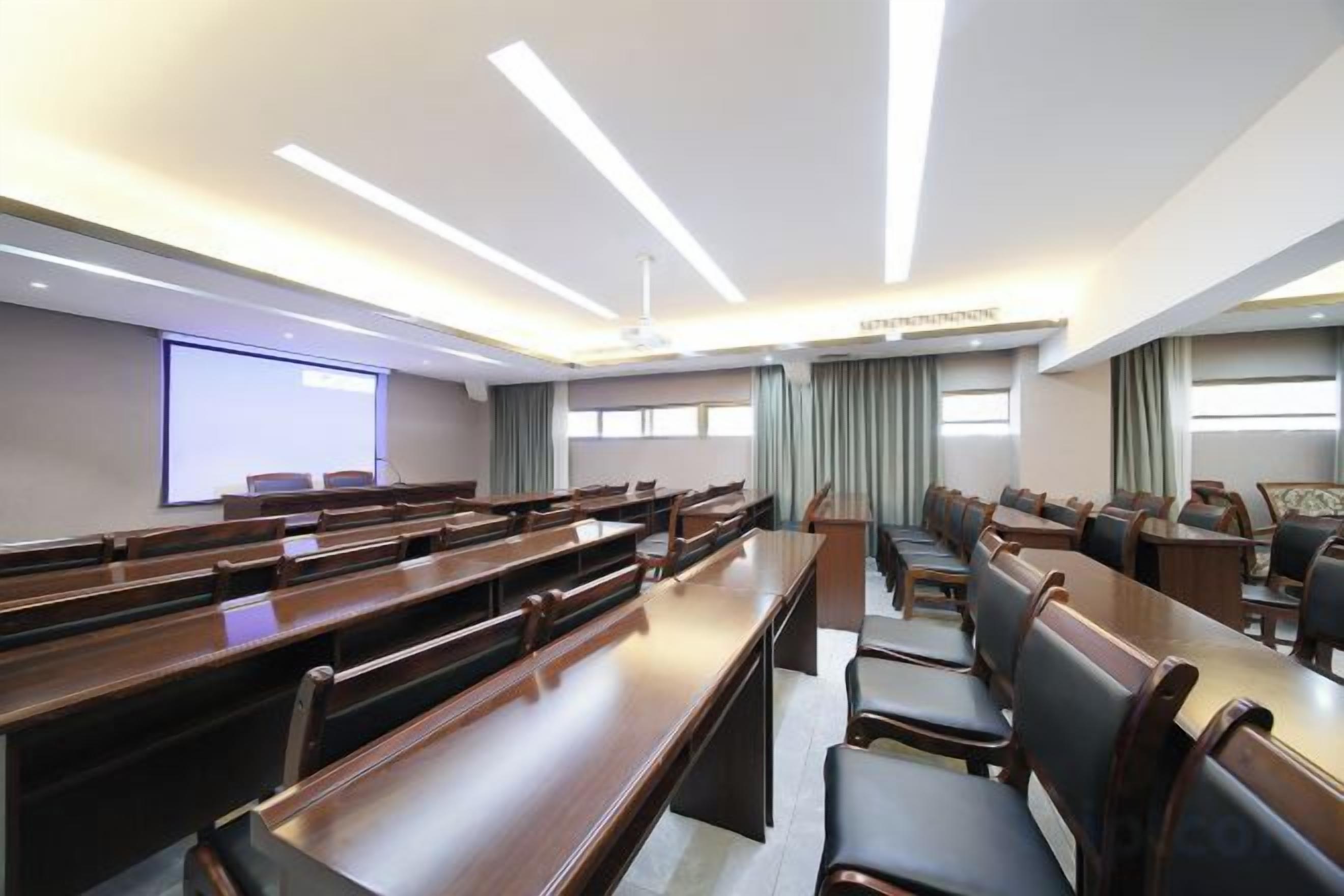 A conference room with rows of wooden tables and chairs, a projector screen, and a large window with curtains.