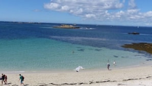Une plage à l’eau turquoise et limpide, avec un rivage sablonneux, où quelques personnes se promènent.
