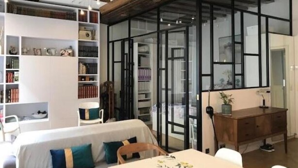 A modern living room with a sofa, a dining table, and a wooden desk. There are bookshelves and a glass partition. - Le Marais Historique rue des Rosiers (Paris)