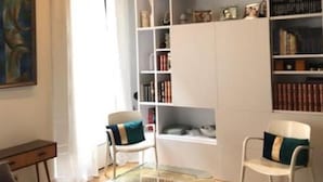 A modern living room with a glass coffee table, two white chairs with teal cushions, a bookshelf filled with books and decorative items, and a painting on the wall. - Le Marais Historique rue des Rosiers (Paris)