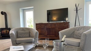 A living room with two grey sofas, a wooden coffee table with a tray of glasses and a champagne bucket, a TV on a wooden cabinet, and a fireplace. - Gîte 7 people -  2 swimming pools (1 with telescopic dome) - The beach on foot (Trégunc)
