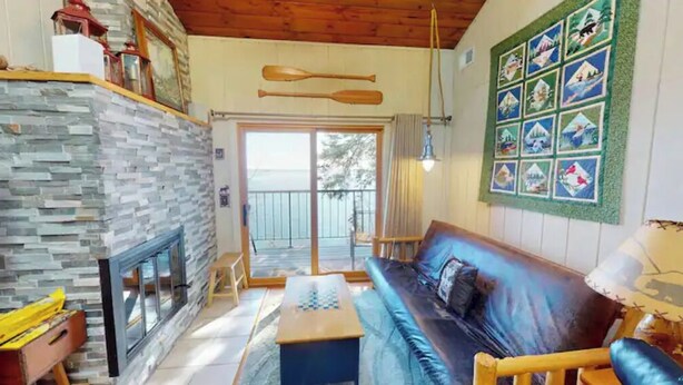 A cozy living room with a stone fireplace, a blue sofa, and a wooden ceiling. - Cedar’s Seaside Chalet (Tofte)