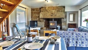 A cozy living room with a stone fireplace, a television, and a dining area with a striped tablecloth. - Gîte 4 people -  2 swimming pools (1 with telescopic dome) - The beach on foot (Trégunc)