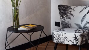 A room with a wooden floor, a black metal side table with a glass vase containing yellow flowers, a black and white patterned armchair, and a plain wall.