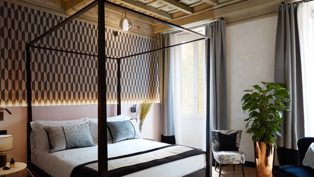A four-poster bed with a geometric patterned headboard, a bedside table with a lamp, a potted plant, and a chair with a patterned cushion.