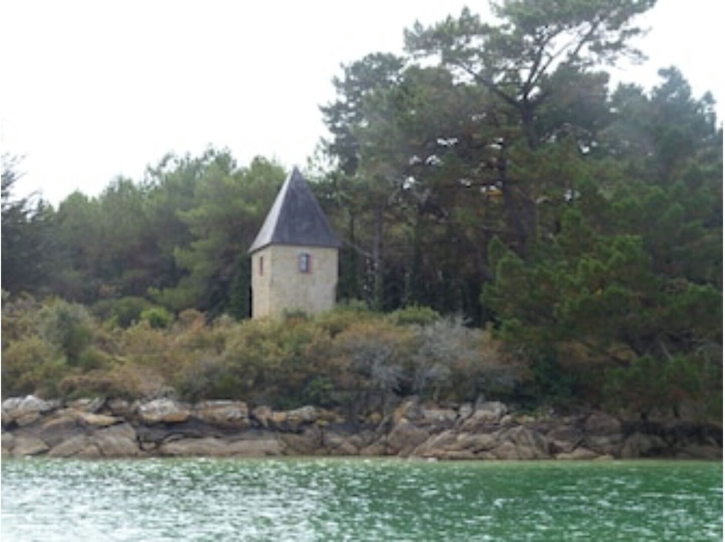 A small stone tower surrounded by dense trees and a rocky shoreline.
