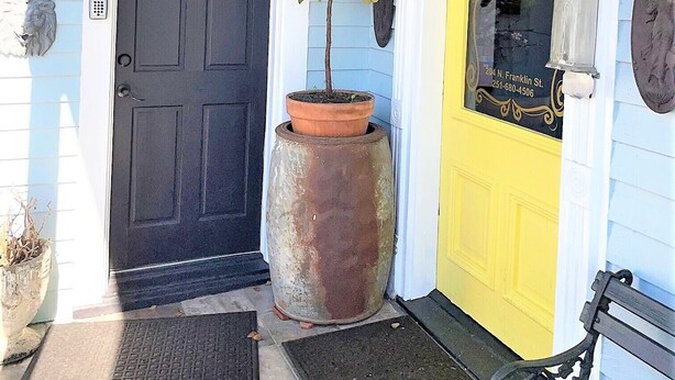 Une porte d’entrée avec une porte noire et une porte jaune, toutes deux portant des numéros au-dessus. Un pot de fleurs se trouve à côté de la porte noire et un banc est placé à droite.