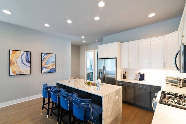 A modern kitchen with a marble island, bar stools, stainless steel appliances, and artwork on the wall.