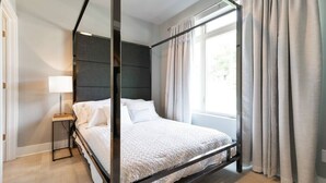 Un lit à baldaquin avec une tête de lit sombre, une table de chevet avec une lampe et des rideaux de couleur claire.
