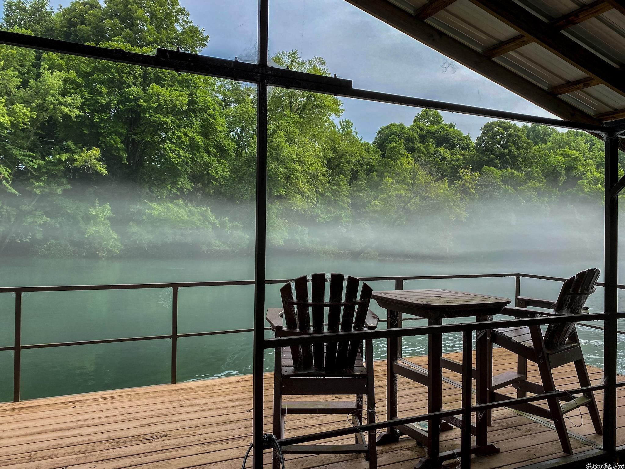 A deck with a table and two Adirondack chairs overlooking a misty river surrounded by dense greenery.