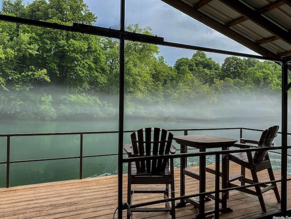 A deck with a table and two Adirondack chairs overlooking a misty river surrounded by dense greenery. - Beautiful House on the River! Book now for Spring & Summer 2025! (Pangburn)