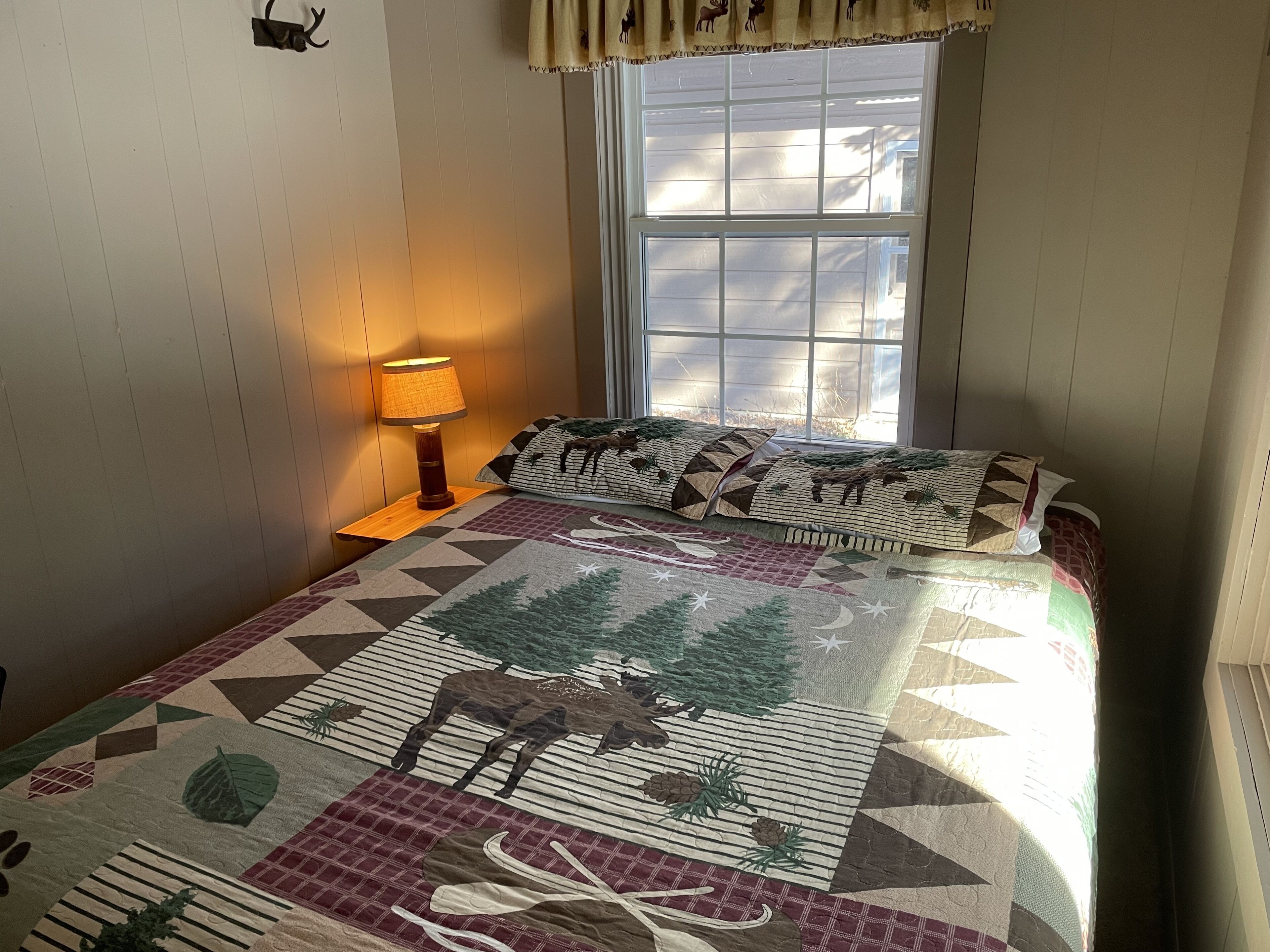 A bedroom with a large window, a bed with a patterned quilt, a bedside lamp, and a wall-mounted clock.