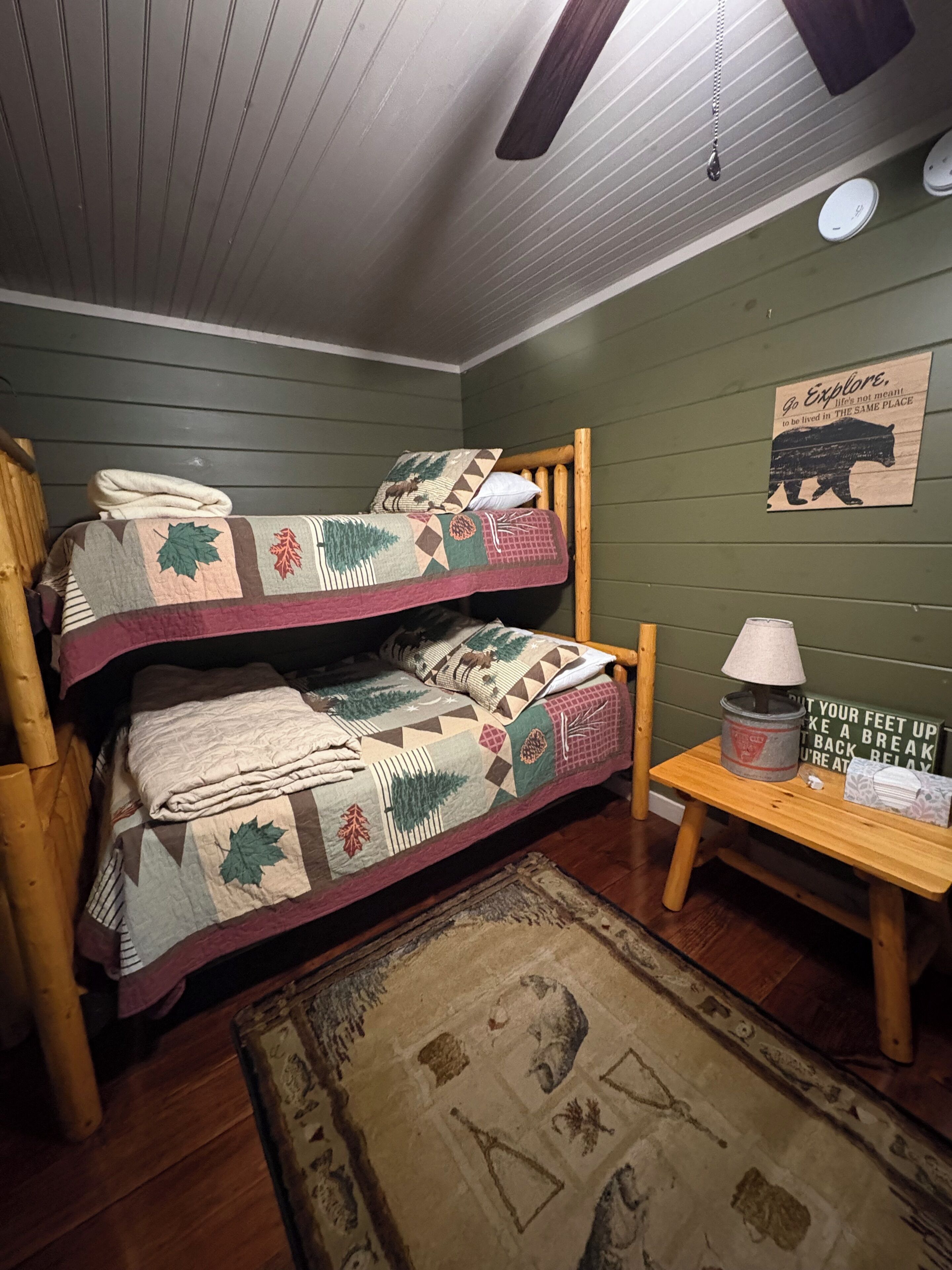 A bunk bed with a quilt, a wooden table with a lamp, and a rug on the floor.