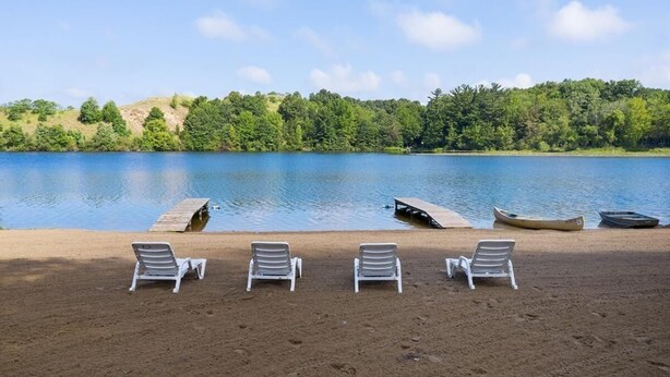 A lakeside setting with four white lounge chairs aligned on a sandy beach, a dock with canoes, and a forested hill in the background. - Sleeps 10-Hot Tub-Close to Downtown-Resort Pool (Saugatuck Township)
