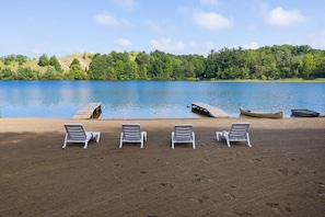 A lakeside setting with four white lounge chairs aligned on a sandy beach, a dock with canoes, and a forested hill in the background.