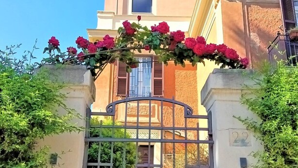 Una entrada protegida con una reja de hierro forjado, un balcón con rosas rojas y un edificio de fachada color crema y contraventanas.