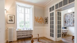 Una habitación con un ventanal grande, un radiador, un zapatero, una escultura de madera y una alfombra estampada.