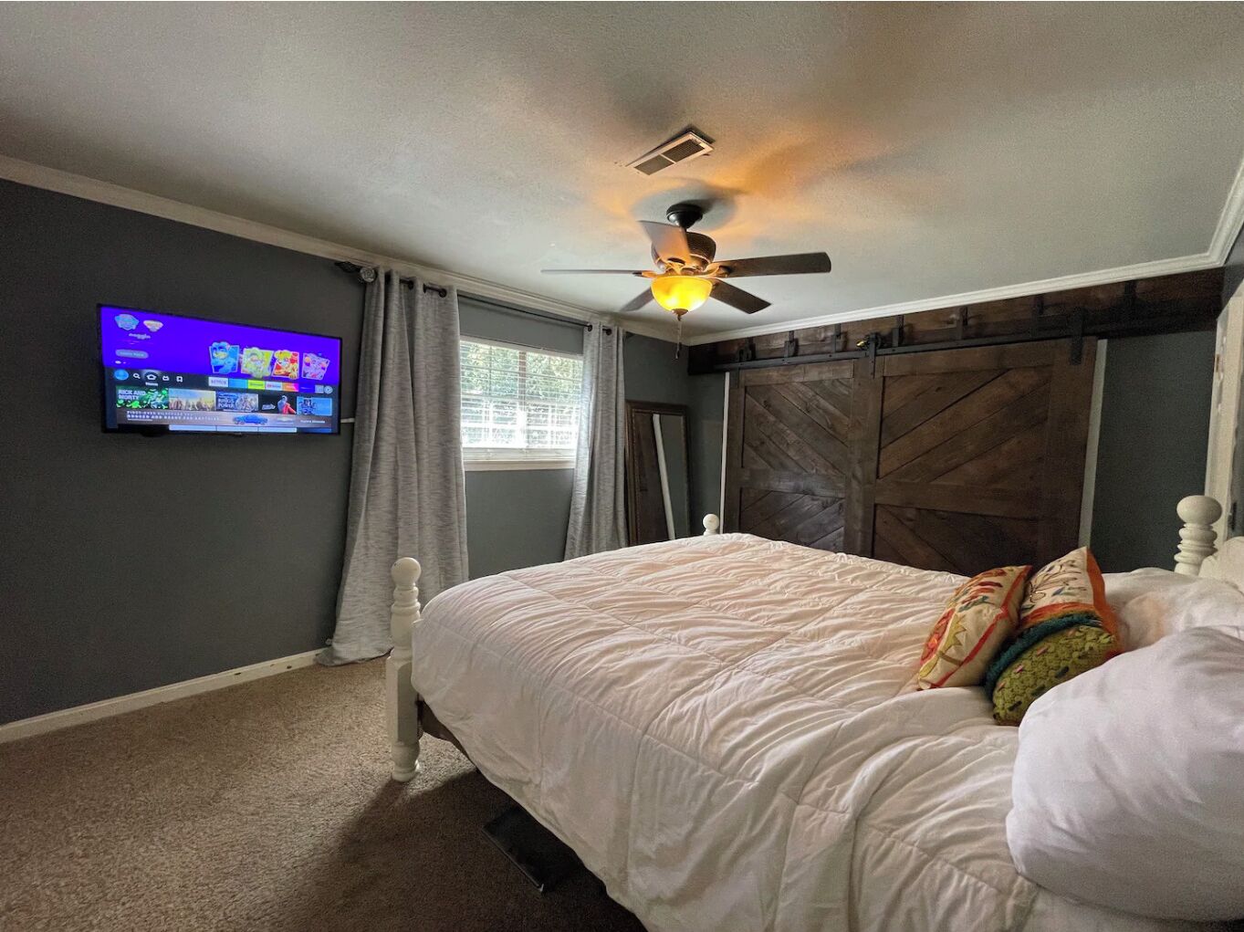 A bedroom with a bed, a ceiling fan, a TV mounted on the wall, and a window with curtains.