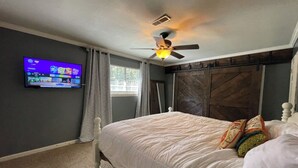 Ein Schlafzimmer mit einem Bett, einem Deckenventilator, einem an der Wand befestigten Fernseher und einem Fenster mit Vorhängen.