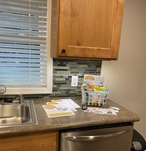 A kitchen with a stainless steel sink, a dishwasher, wooden cabinets, a window with blinds, and a countertop with a basket of snacks and check-in/out papers. - Boomerang Home Rentals - # 4au-L (Janesville)