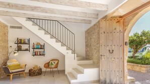 A modern interior with a staircase, bookshelves, a sofa, and a wooden door leading outside. - Can Corme (Pollensa)