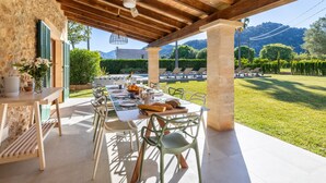 A covered outdoor dining area with a table set for four, chairs, and a view of a lush garden and mountains in the background. - Can Corme (Pollensa)