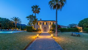 A stone building with a paved pathway leading to a wooden door, surrounded by palm trees and a pool. - Can Corme (Pollensa)