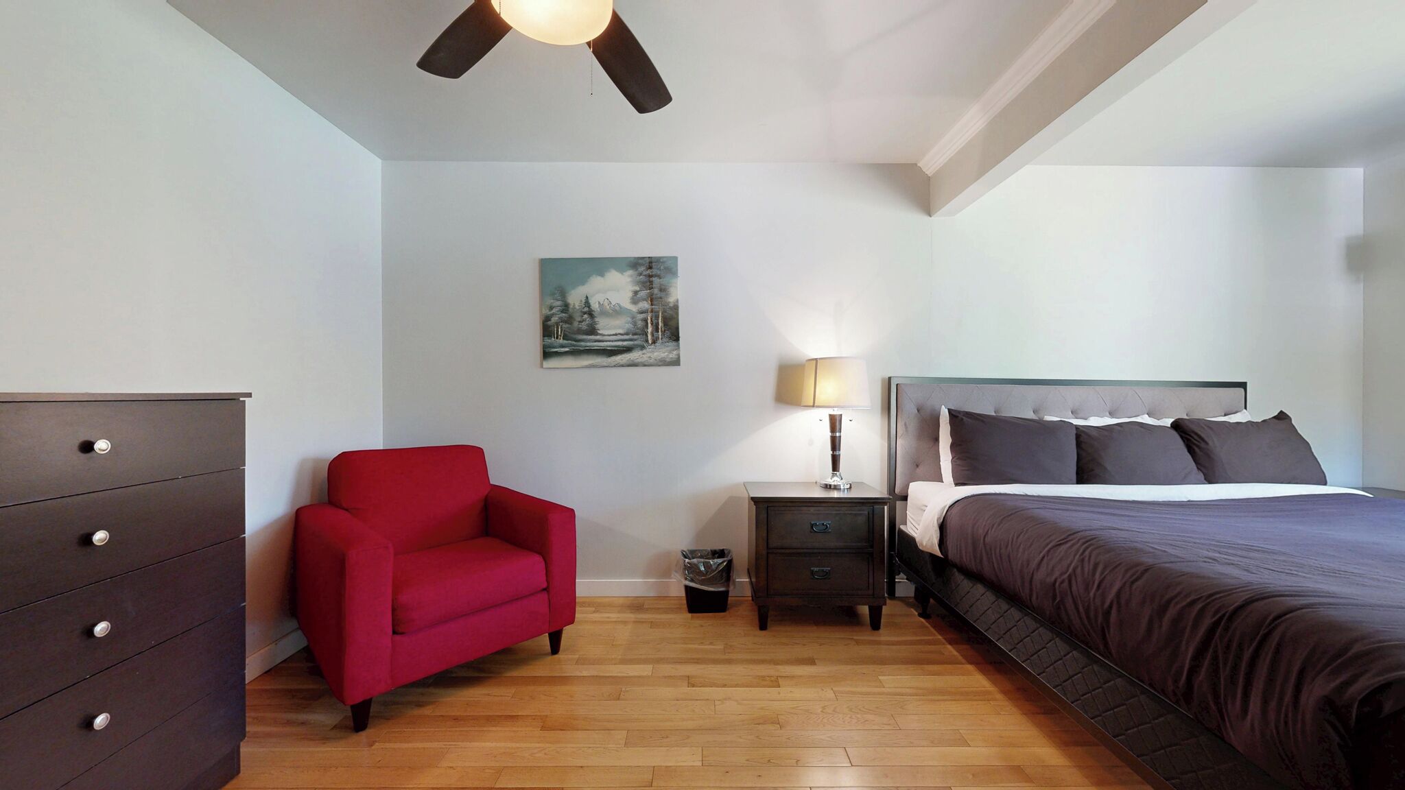 A bedroom with a bed, a red chair, a dresser, a nightstand with a lamp, and a painting on the wall.