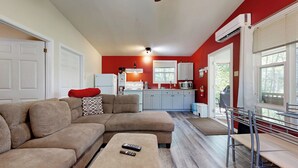 Una sala moderna con un sofá modular, una mesita con controles remotos, una cocina con armarios blancos y una pared de acento roja.