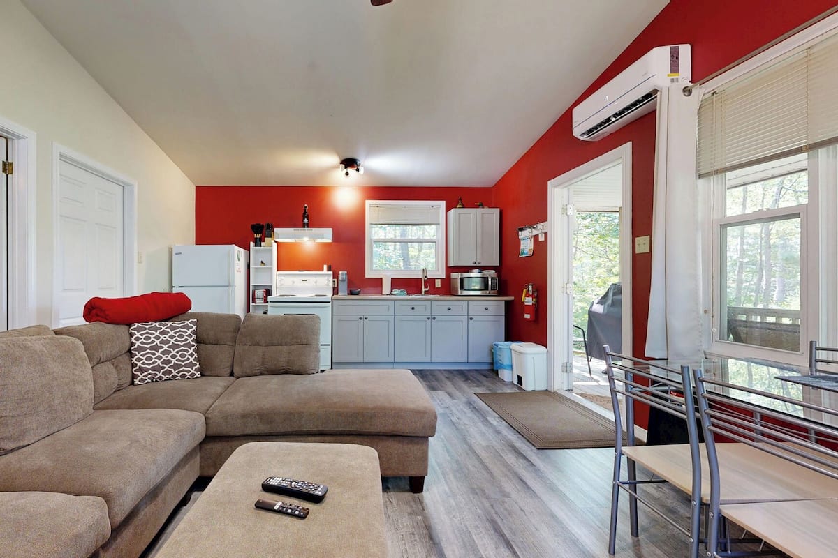 Una sala moderna con un sofá modular, una mesita con controles remotos, una cocina con armarios blancos y una pared de acento roja.