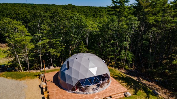 Una estructura de cúpula geodésica con terraza de madera y escalones de piedra, rodeada de zona boscosa.