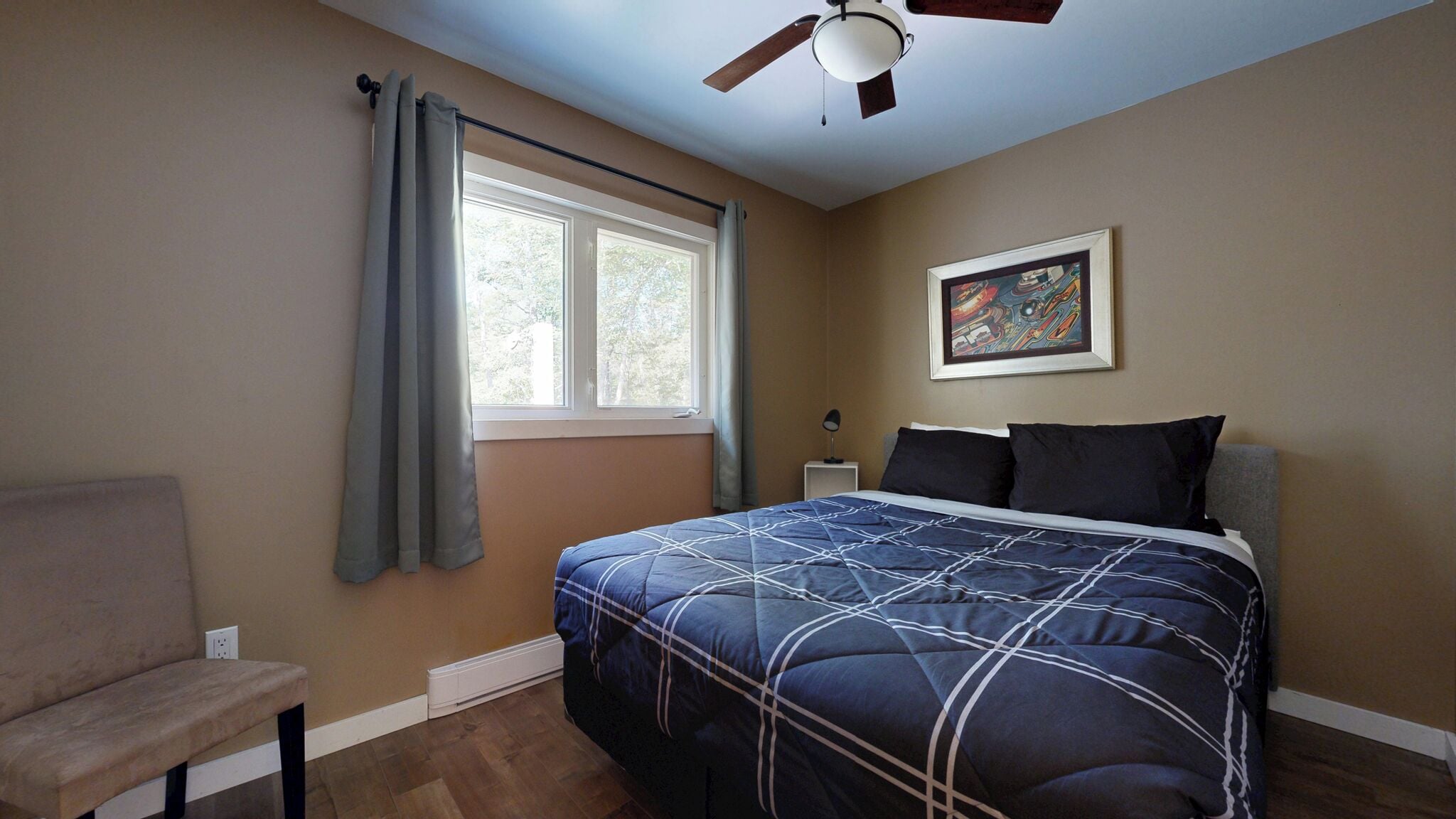Quarto com cama, uma cadeira, ventoinha no teto, janela com cortinas, mesa de cabeceira e um quadro na parede.
