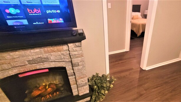 A living room with a stone fireplace, a flat-screen TV displaying streaming services, and a hallway leading to a bedroom. - Super Cozy Home Away From Home (Abilene)
