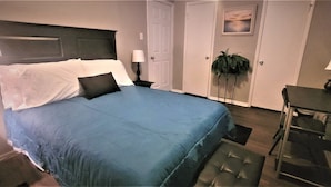 A neatly made bed with a blue bedspread and white pillows, a bedside table with a lamp, a small potted plant, and a desk with a lamp. - Super Cozy Home Away From Home (Abilene)