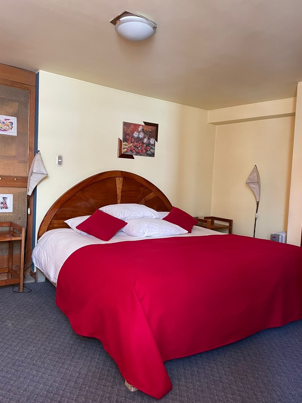 Habitación de hotel con cama de madera, ropa de cama roja, una mesita de noche, una lámpara y un cuadro colgado en la pared.