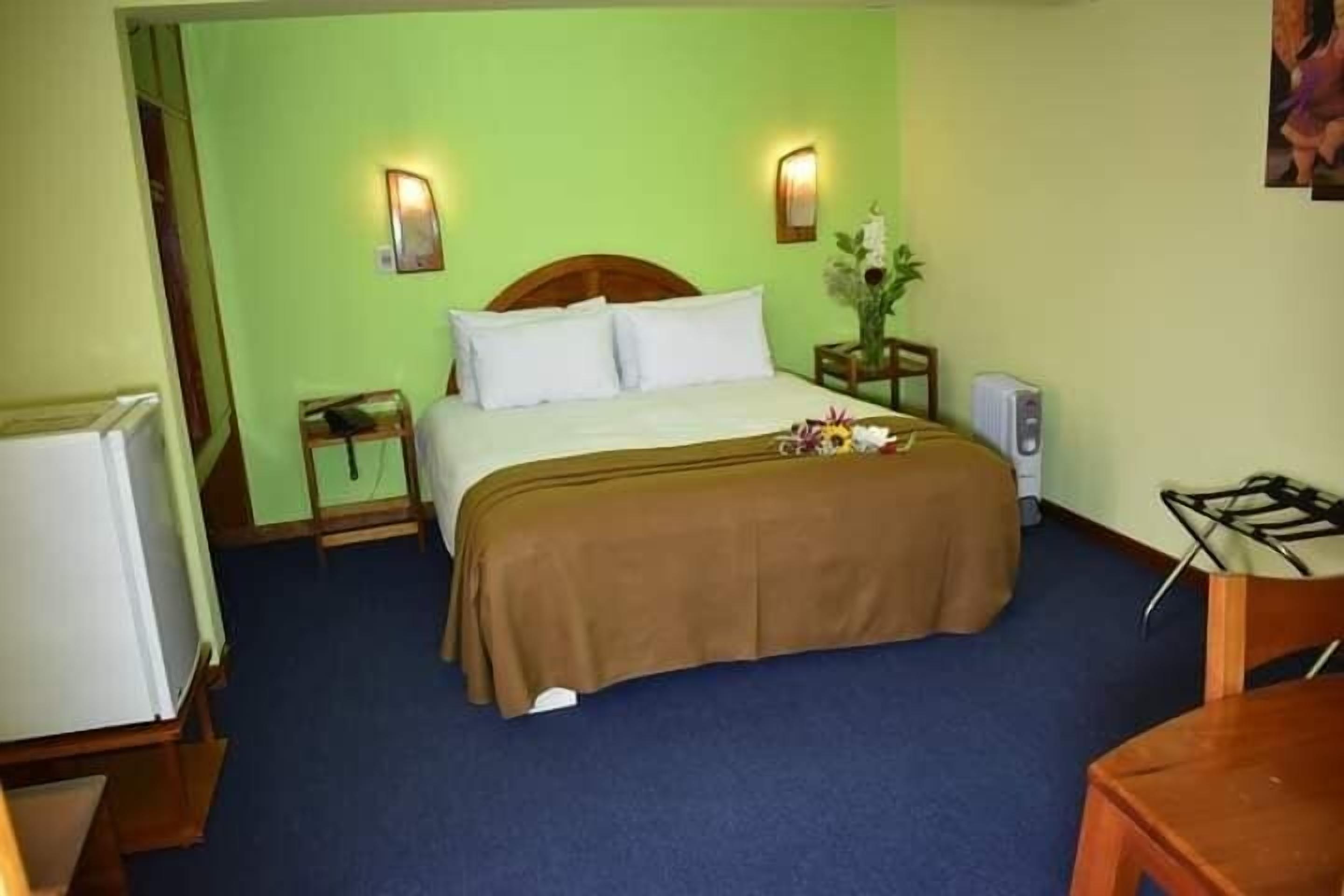 A hotel room with a bed, a nightstand, a bedside table, a chair, a desk, a lamp, a wall-mounted TV, and a wall-mounted light fixture.