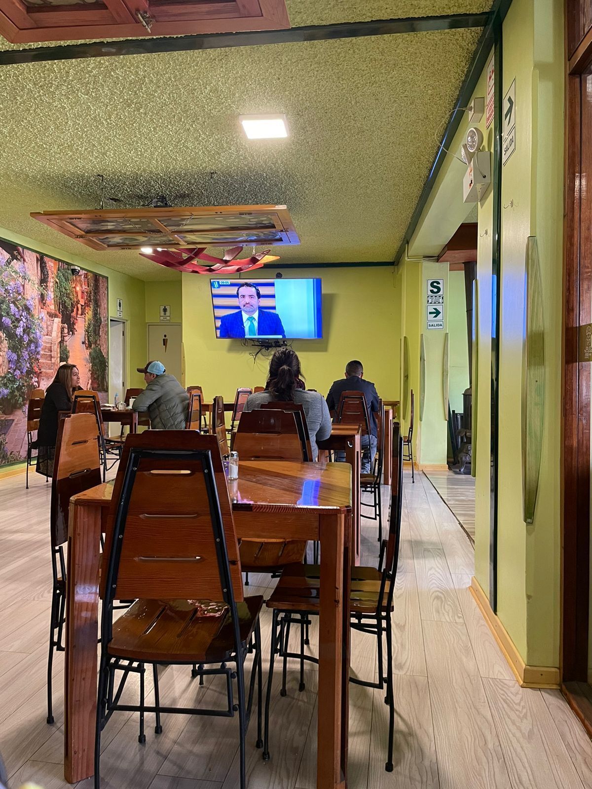 Un restaurante con mesas y sillas de madera, una pantalla de televisión mostrando un noticiero y un mural en la pared.