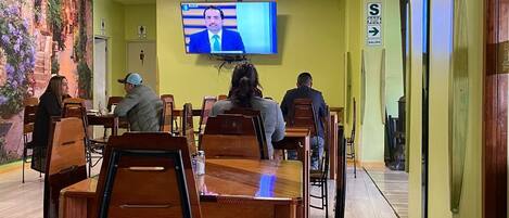 Un restaurante con mesas y sillas de madera, una pantalla de televisión mostrando un noticiero y un mural en la pared.