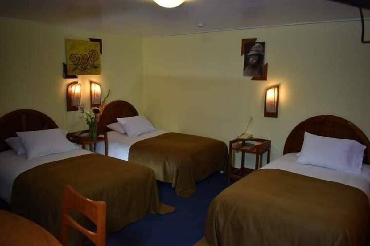 A hotel room with three single beds, each with a wooden headboard and a brown bedspread. There are wall-mounted lamps on either side of the beds.