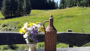 Une table en bois rustique, ornée d’un vase de fleurs, d’une bouteille en osier et de deux verres à liqueur.
