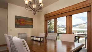 A dining room with a wooden table, six chairs, a chandelier, and a large window with a view of a snowy landscape. - Majestic Mountain Family Escape - 4 bedroom/4Bathroom with lots of room and easy access to the slopes, river and town (Steamboat Springs)