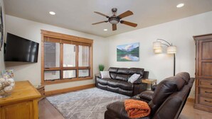 A living room with a brown leather sofa, a wooden coffee table, a TV mounted on the wall, a ceiling fan, and a large window with blinds. - Majestic Mountain Family Escape - 4 bedroom/4Bathroom with lots of room and easy access to the slopes, river and town (Steamboat Springs)