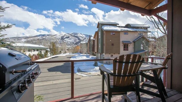 A wooden deck with a grill and a chair, overlooking a snowy landscape and a building with a green roof. - Majestic Mountain Family Escape - 4 bedroom/4Bathroom with lots of room and easy access to the slopes, river and town (Steamboat Springs)
