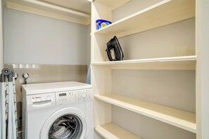 A white Bosch washing machine in a laundry room with a shelf above it and a blue container labeled 'Cold Power' on the top shelf. - 1BR unit in the heart of Manly (Manly)