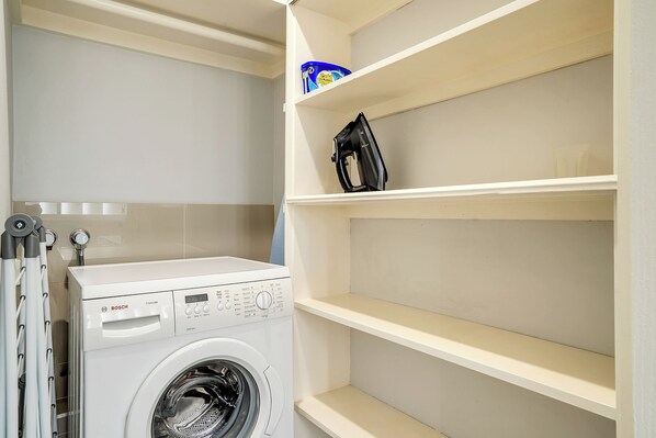 Una lavadora blanca de Bosch en un cuarto de lavado, con un estante encima y un recipiente azul etiquetado 'Cold Power' en la parte superior.