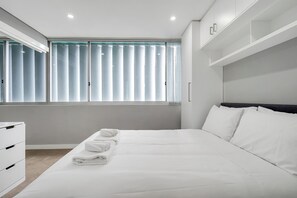 Habitación de hotel moderna con una cama grande, una mesita de noche y un ventanal con persianas.