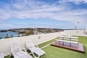 A rooftop terrace with white lounge chairs, a wooden deck, and a view of a coastal town. - 1BR unit in the heart of Manly (Manly)