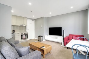 A modern living room with a sofa, coffee table, TV, and a small kitchen area with cabinets and appliances. - 1BR unit in the heart of Manly (Manly)
