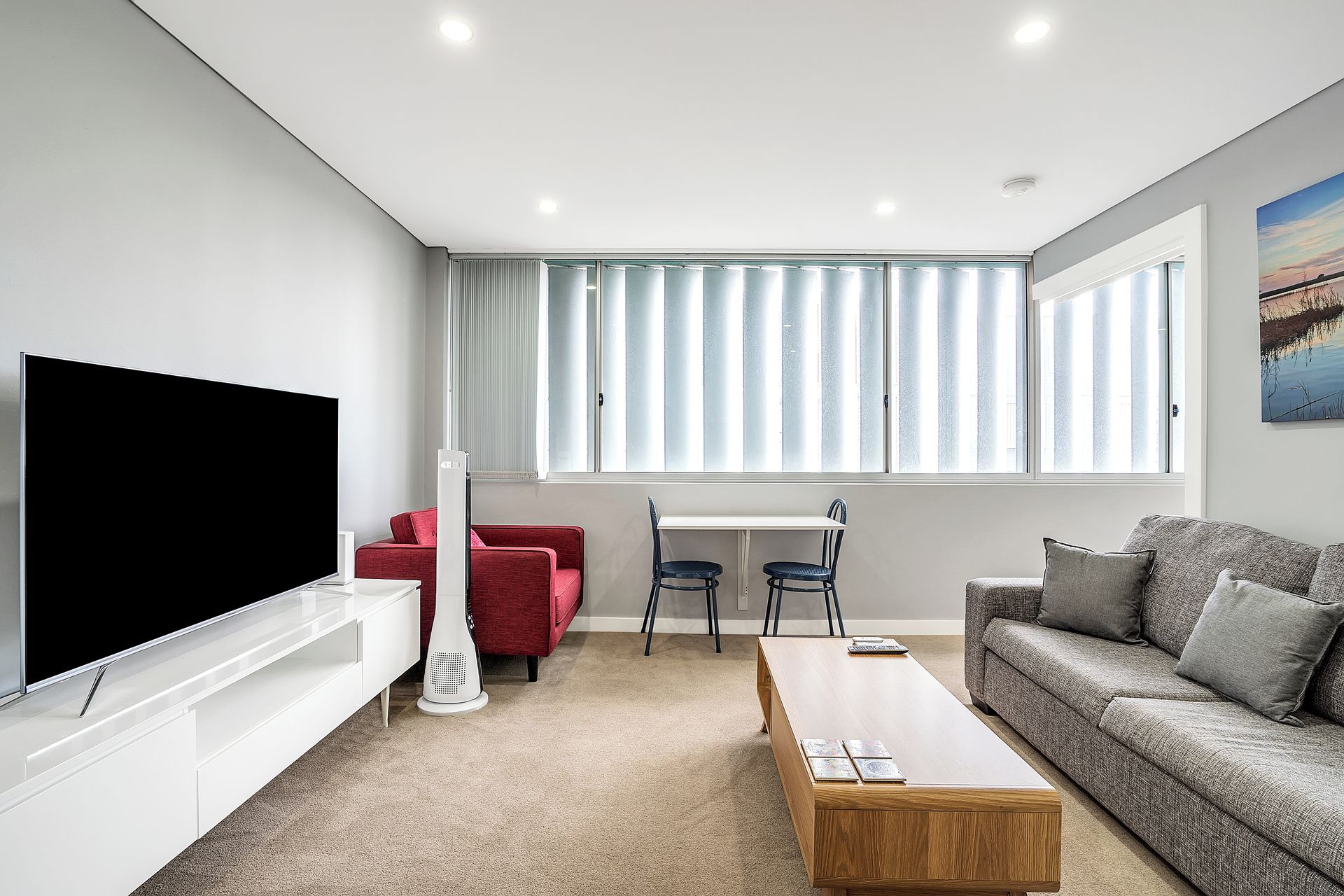 A modern living room with a flat-screen TV, a red sofa, a grey sofa, a wooden coffee table, and a dining area with a white table and two chairs.