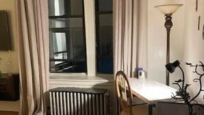A room with a wooden floor, a radiator, a table with a chair, a lamp, and a window with curtains. - PLG Boho Stay (Flatbush) (Brooklyn)